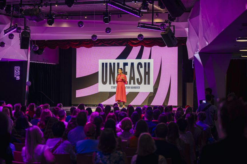 Frau in rotem Kleid spricht auf Bühne vor Publikum, hinter ihr eine Leinwand mit der Aufschrift „UNLEASH".