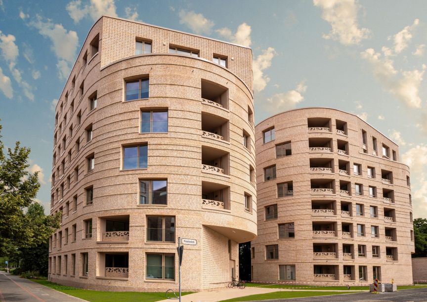 Modernes mehrstöckiges Wohngebäude aus hellem Backstein mit geschwungener Fassade, Balkonen und großen Fenstern, blauer Himmel.
