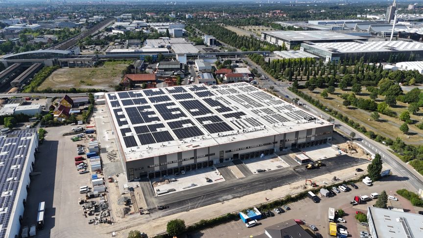 Luftaufnahme eines großen Industriegebäudes mit Solaranlage auf dem Flachdach, umgeben von Industriegebiet und Grünflächen.