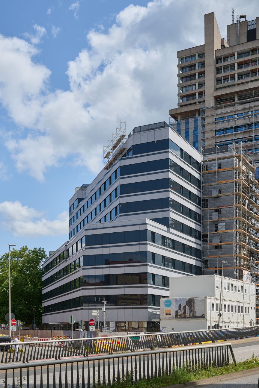 Modernes Bürogebäude mit blau-weißer Fassade neben einem Hochhaus mit Baugerüst, davor Straße und Zaun.