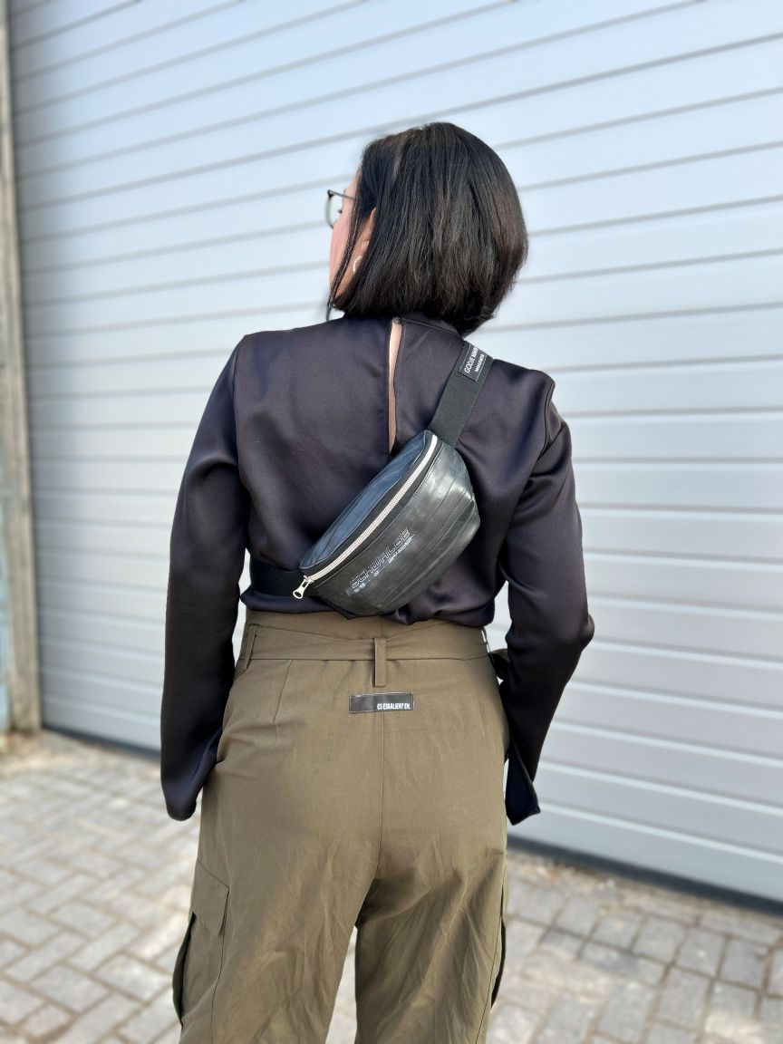 Person von hinten, schwarze Bluse, olivgrüne Cargohose, schwarze Gürteltasche quer getragen, dunkle Haare, Brille.