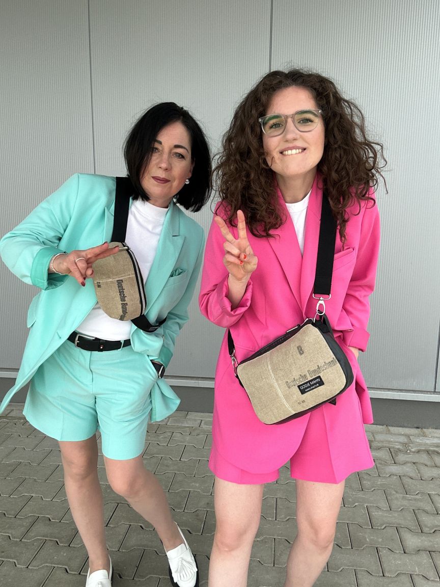 Zwei Frauen posieren lächelnd: eine im türkisen, die andere im pinken Blazer-Shorts-Outfit, beide mit beigen Umhängetaschen.
