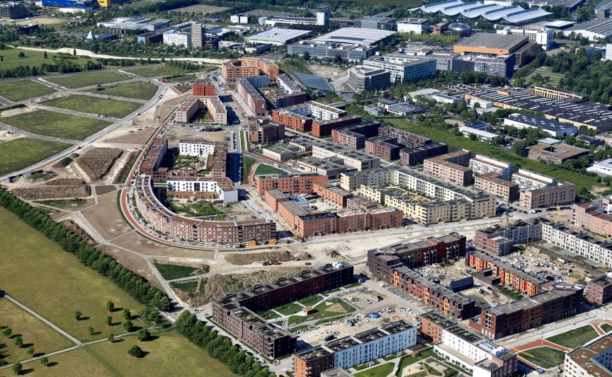 Luftaufnahme einer Stadtentwicklung mit mehrstöckigen Wohngebäuden, Baustellen, Grünflächen und Industriegebäuden im Hintergrund.