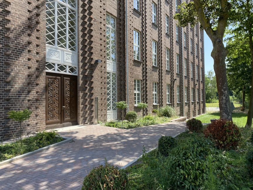 Mehrstöckiges Backsteingebäude mit verzierten Holztüren, Sprossenfenstern und gepflastertem Gehweg, umgeben von Bäumen und Sträuchern.