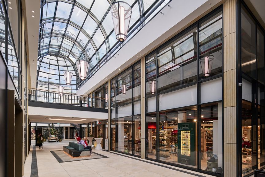 Helles Einkaufszentrum mit Glasdach, eleganten Hängelampen, zwei Etagen, Geschäften hinter Glasfronten und Sitzmöbeln im Gang.