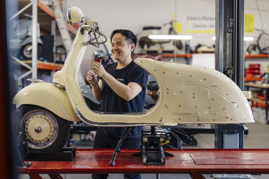 Mann in schwarzem T-Shirt arbeitet in Werkstatt an cremefarbenem Vintage-Roller auf Hebebühne.