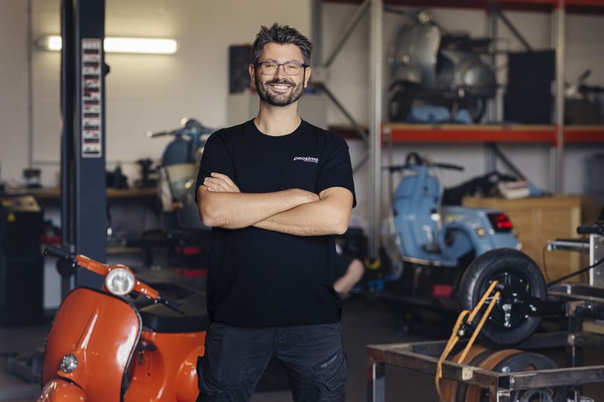 Mann mit Brille und schwarzem T-Shirt steht mit verschränkten Armen in einer Werkstatt mit Motorrollern.