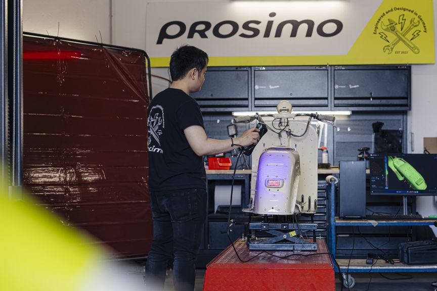 Ein Mann in schwarzem T-Shirt arbeitet in einer Werkstatt an einem Roller. Im Hintergrund das Logo „Prosimo".