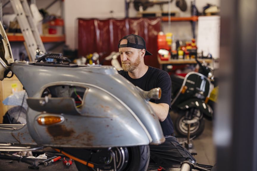 Mann mit Bart und Basecap repariert ein graues Motorrad in einer Werkstatt. Im Hintergrund weitere Fahrzeuge.
