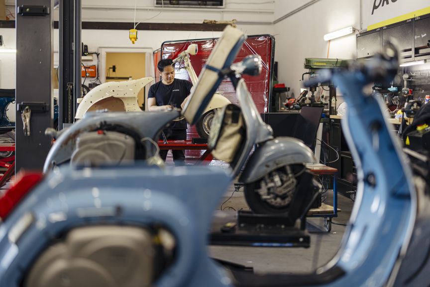 Ein Mechaniker arbeitet in einer Werkstatt zwischen mehreren blauen und roten Motorrollern in verschiedenen Zerlegungszuständen.
