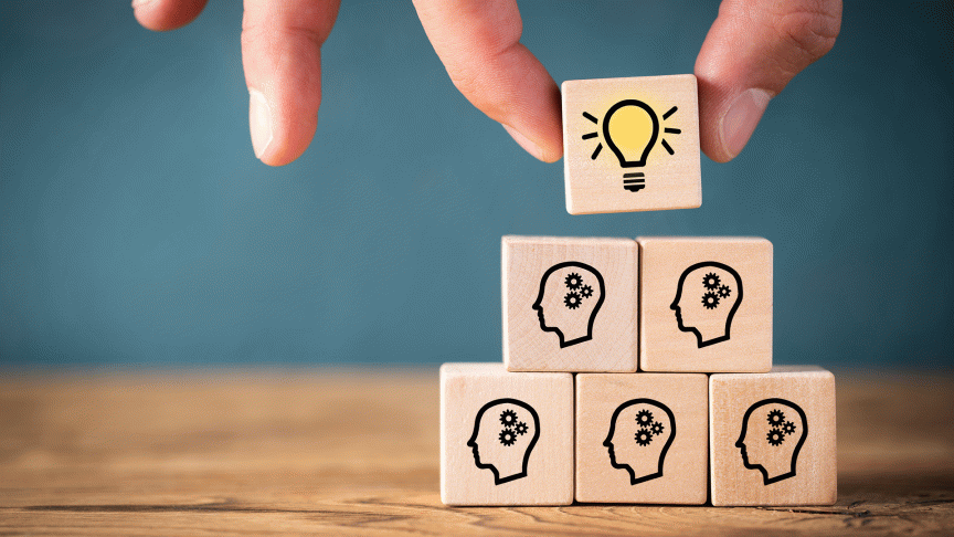 Sechs Holzwürfel als Pyramide, fünf davon mit Köpfen und Zahnräder und der oberste Würfel mit Glühbirne.