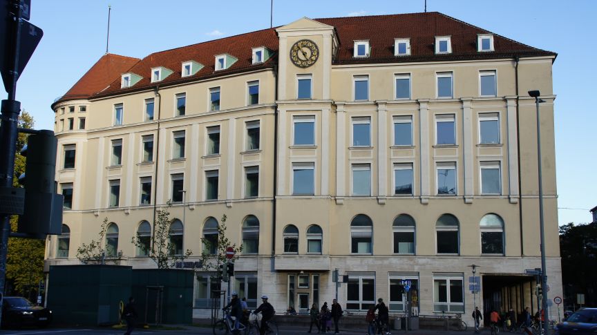 Mehrstöckiges Gebäude mit cremefarbener Fassade, rotem Dach und Uhr, belebt von Fußgängern an einer Straßenecke.