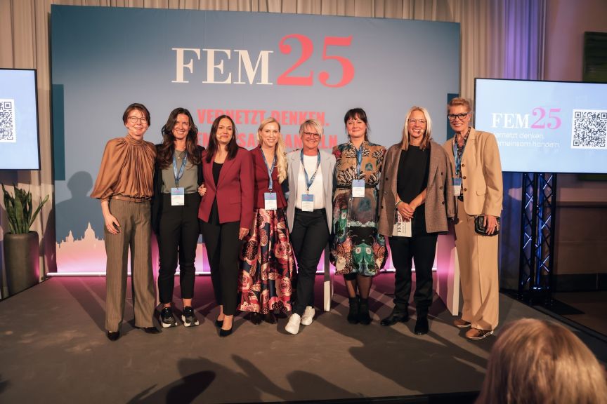 Acht Frauen stehen lächelnd auf einer Bühne vor einem blauen Banner mit der Aufschrift „FEM25".