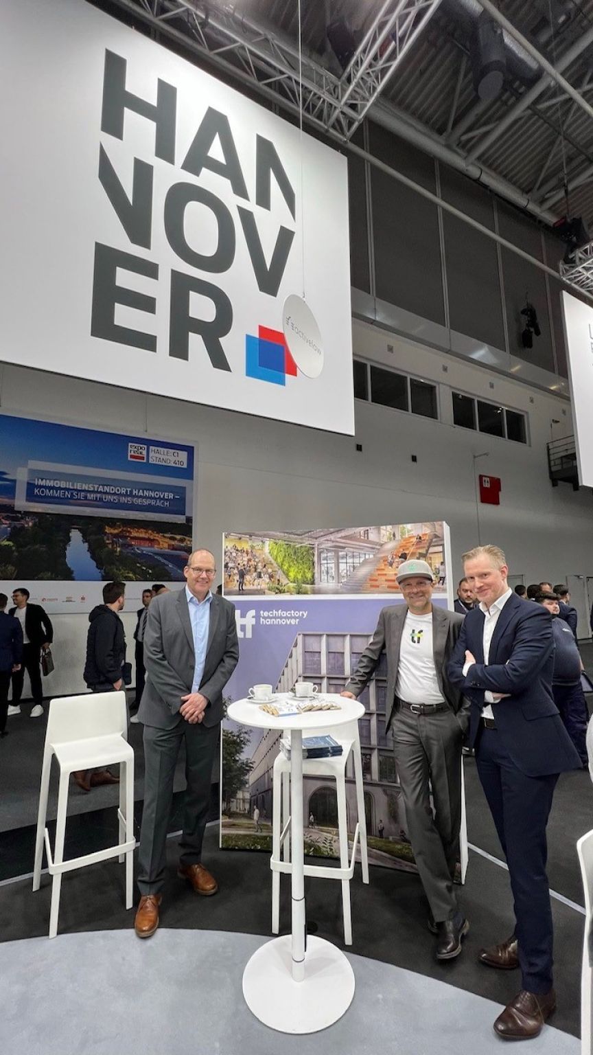 Drei Männer stehen am Stand der Stadt Hannover auf einer Messe vor einem großen Hannover-Logo.