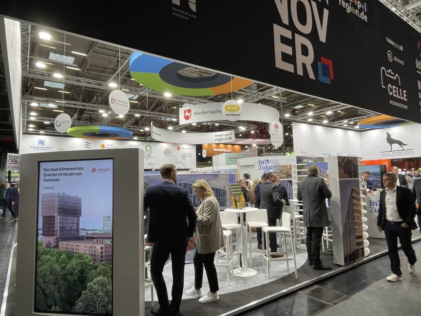 Messestand „Hannover", mehrere Personen im Gespräch, LED-Bildschirm am linken Rand.