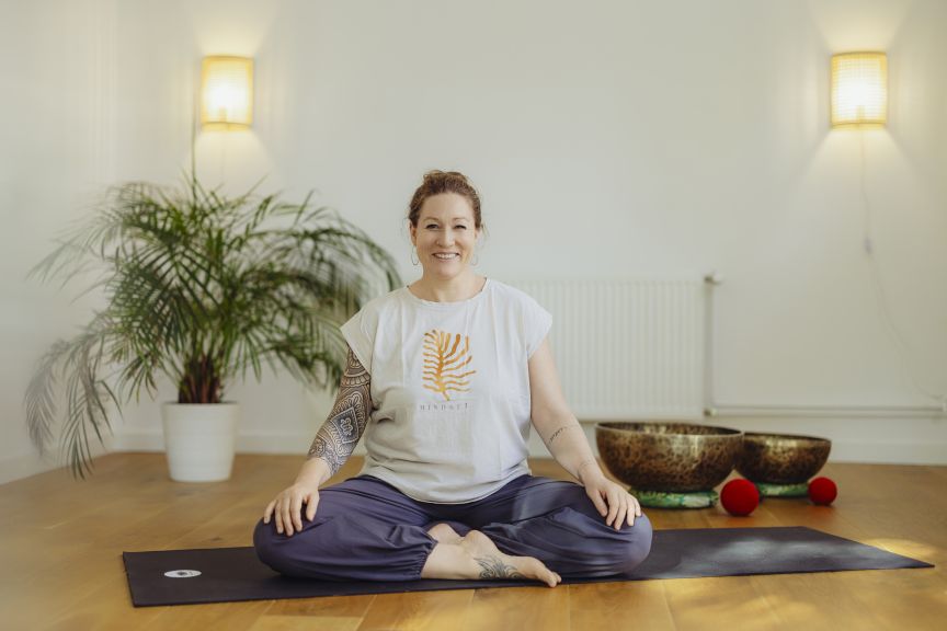 Frau sitzt im Schneidersitz auf Yogamatte, lächelt, trägt weißes T-Shirt, hat Tattoos, Klangschalen im Hintergrund.