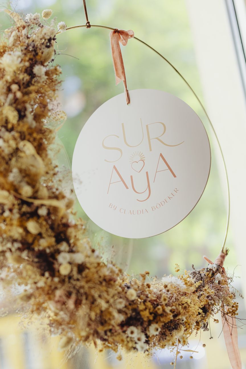 Rundes Schild mit Aufschrift „Suraya by Claudia Bodener" hängt an einem Trockenblumenkranz vor einem Fenster.