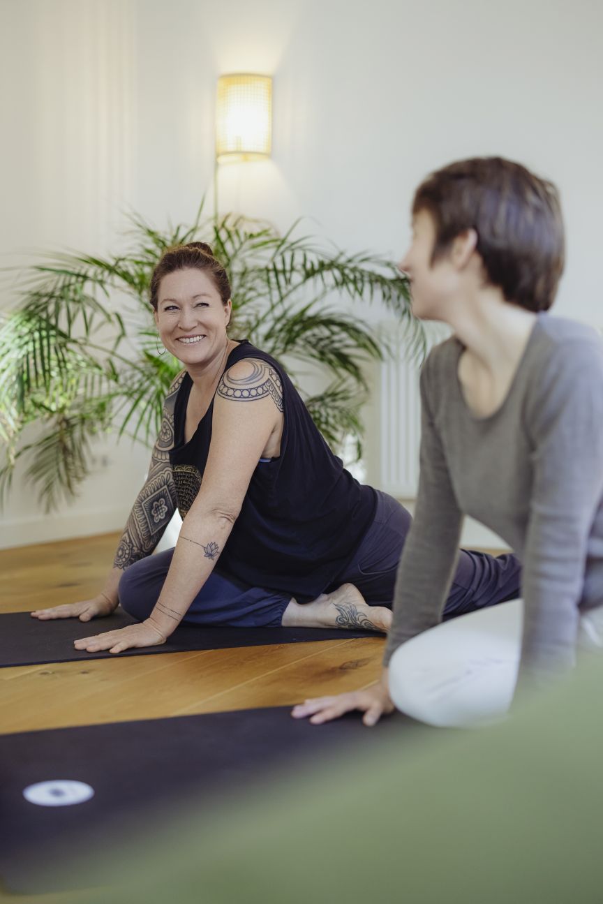 Zwei Frauen auf Yogamatten in hellem Raum mit Pflanze. Eine Frau mit Tattoos lächelt, beide sitzen auf dem Boden.