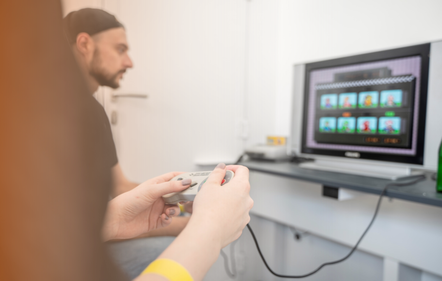 Person hält SNES-Controller, im Hintergrund sitzt ein Mann vor einem Fernseher mit einem Videospiel.