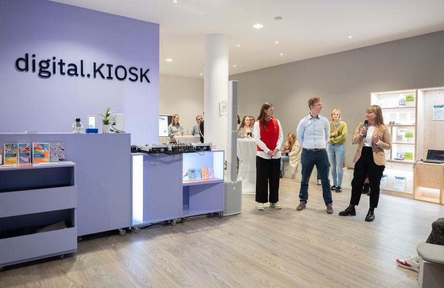 Innenraum eines „digital.KIOSK"-Ladens mit mehreren Personen, beleuchtetem Tresen und Produktregalen im Hintergrund.
