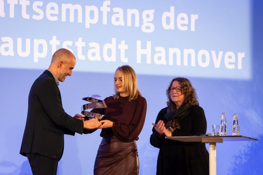 Drei Personen bei einer Preisverleihung in Hannover. Ein Mann überreicht einer Frau eine Auszeichnung, eine zweite Frau applaudiert.