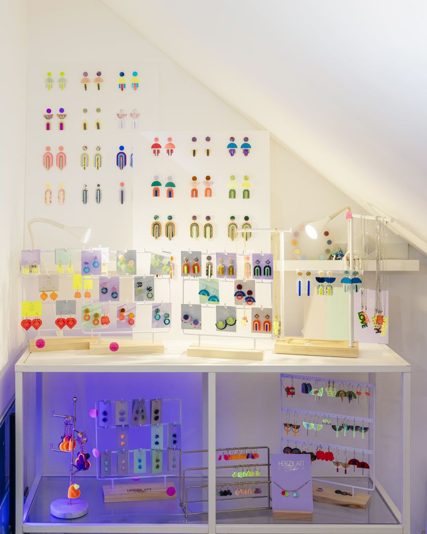 Weißes Regal mit bunten Ohrringen auf Karten, darüber eine Wand mit aufgehängten Ohrringen-Paaren in verschiedenen Farben.