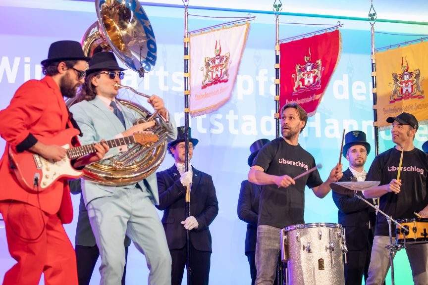 Musiker auf einer Bühne spielen Gitarre, Tuba und Schlagzeug. Im Hintergrund stehen Männer mit Fahnen.