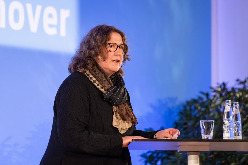 Frau mit Brille und Schal spricht an einem Stehtisch vor blauem Hintergrund mit der Aufschrift „…hover".