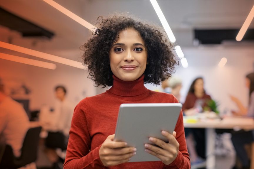 Frau mit lockigem Haar und rotem Rollkragenpullover hält Tablet in einem modernen Büro.