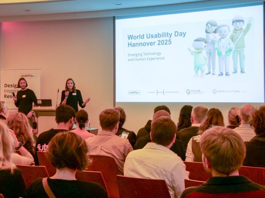 Zwei Personen präsentieren auf einer Bühne. Leinwand zeigt „World Usability Day Hannover 2025". Publikum sitzt im Zuschauerraum.