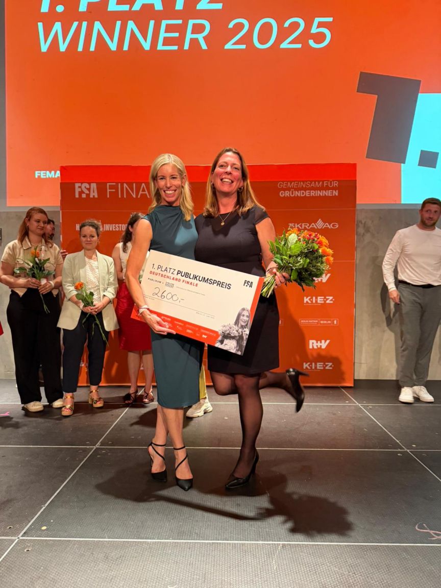 Zwei Frauen auf einer Bühne halten einen Scheck über 2600€ und Blumen. Schriftzug: „1. Platz Winner 2025".
