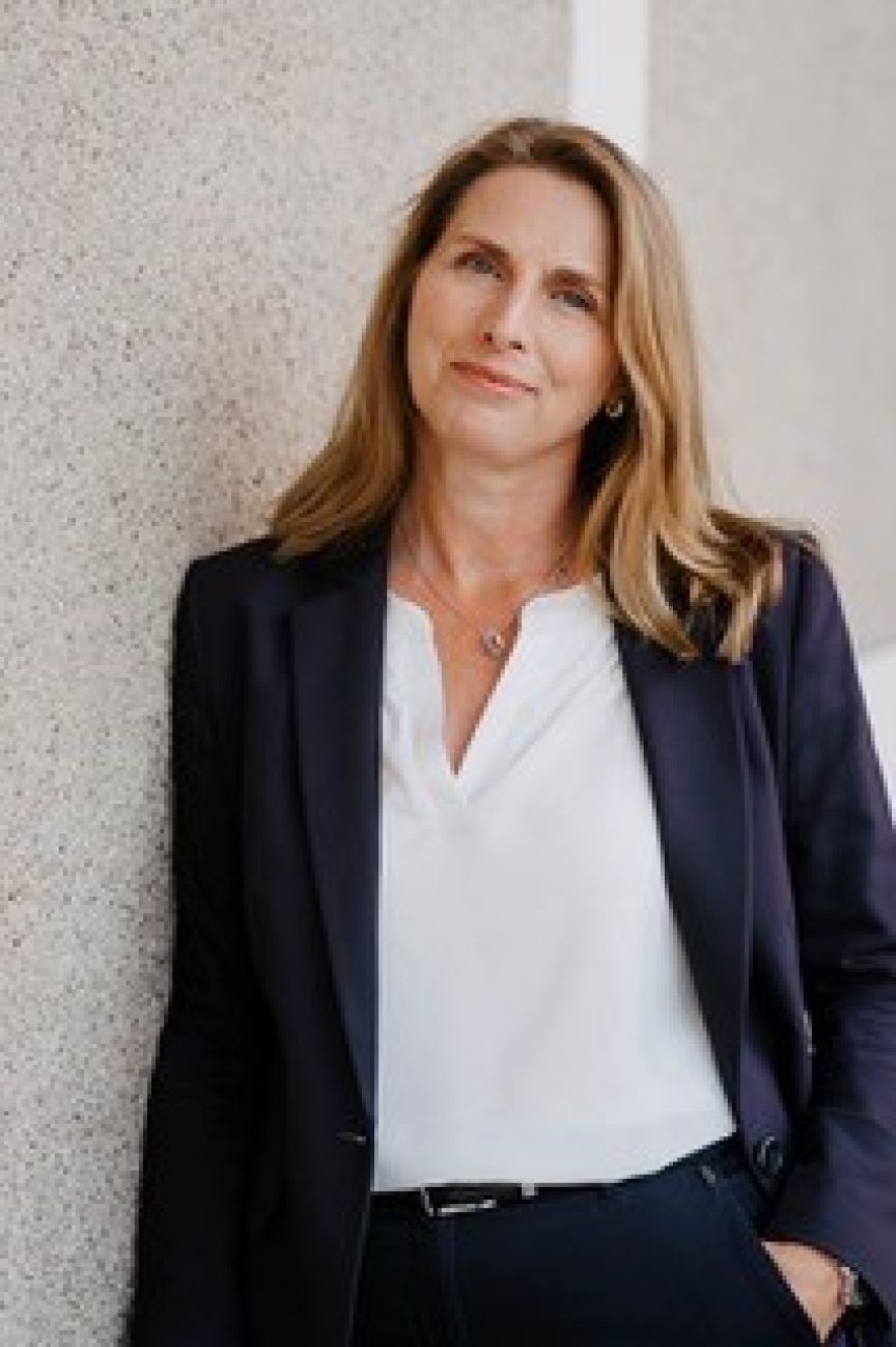 Frau mit langen blonden Haaren, dunklem Blazer und weißer Bluse lehnt an einer hellen Wand.