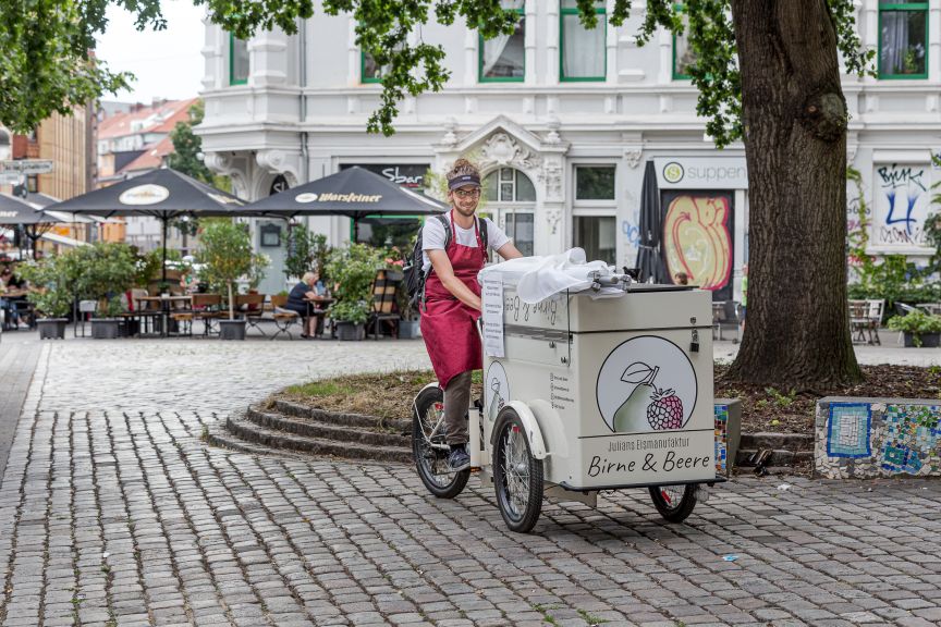Person mit roter Schürze schiebt Lastenrad mit Aufschrift „Birne & Beere" auf einem Kopfsteinpflasterplatz.