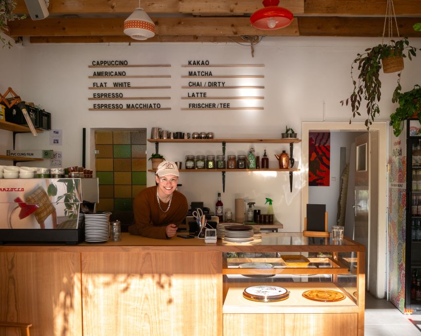 Café-Tresen mit Menütafel, Espressomaschine und Glasvitrine. Eine Person mit Kappe steht lächelnd dahinter.