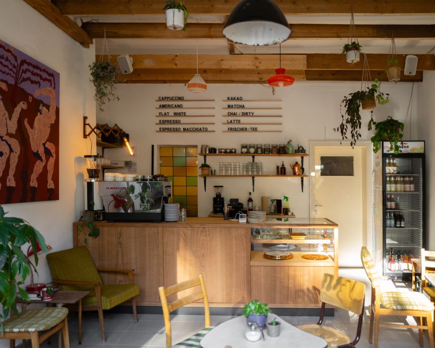 Kleines Café mit Holztresen, Espressomaschine, Speisekarte an der Wand, Hängepflanzen und Sitzgelegenheiten im Vordergrund.