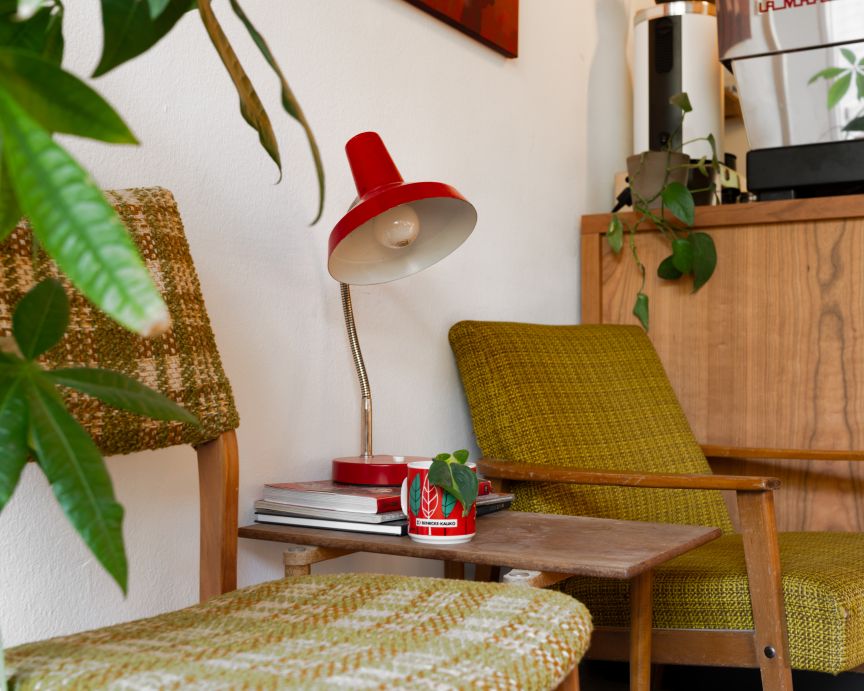 Zwei Retro-Sessel in Olivgrün, ein roter Schreibtischlampe auf Beistelltisch, Zimmerpflanzen, Holzkommode im Hintergrund.