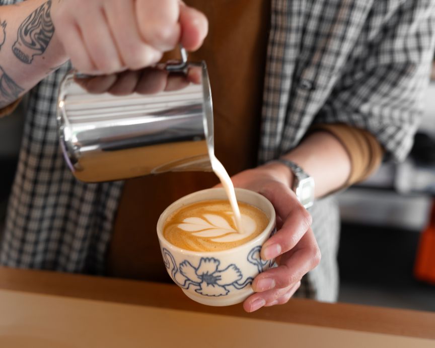 Barista gießt Milchschaum aus Edelstahlkännchen in Tasse mit Blumenmuster, dabei entsteht Latte-Art-Muster.