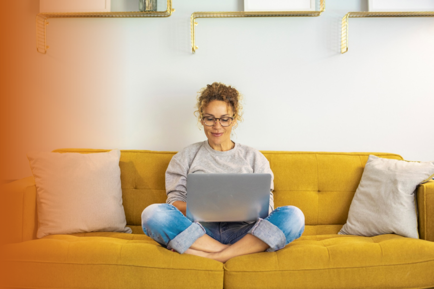 Frau mit Brille sitzt im Schneidersitz auf gelbem Sofa und arbeitet an einem Laptop.