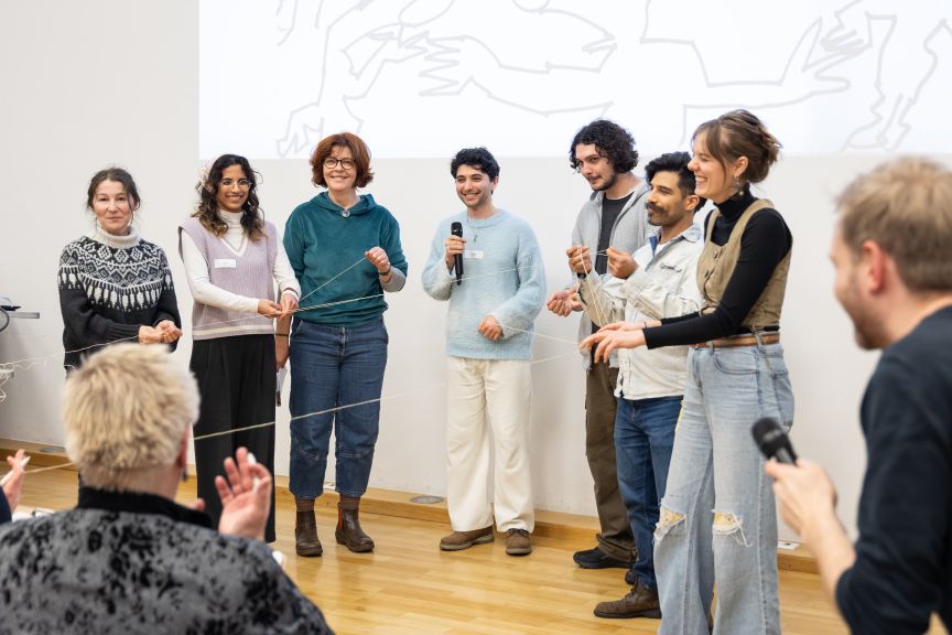 Sieben Personen stehen in einer Reihe vor einer Leinwand und halten gemeinsam ein Seil. Eine Person hält ein Mikrofon.
