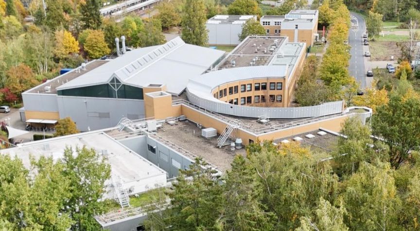 Luftaufnahme eines modernen Gebäudekomplexes mit geschwungener Fassade, umgeben von herbstlichen Laubbäumen.