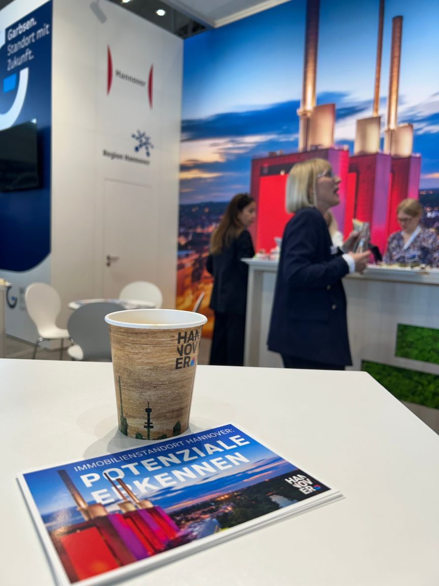 Messestand der Region Hannover mit Kaffeebecher und Broschüre „Potenziale erkennen" im Vordergrund, zwei Frauen im Gespräch.