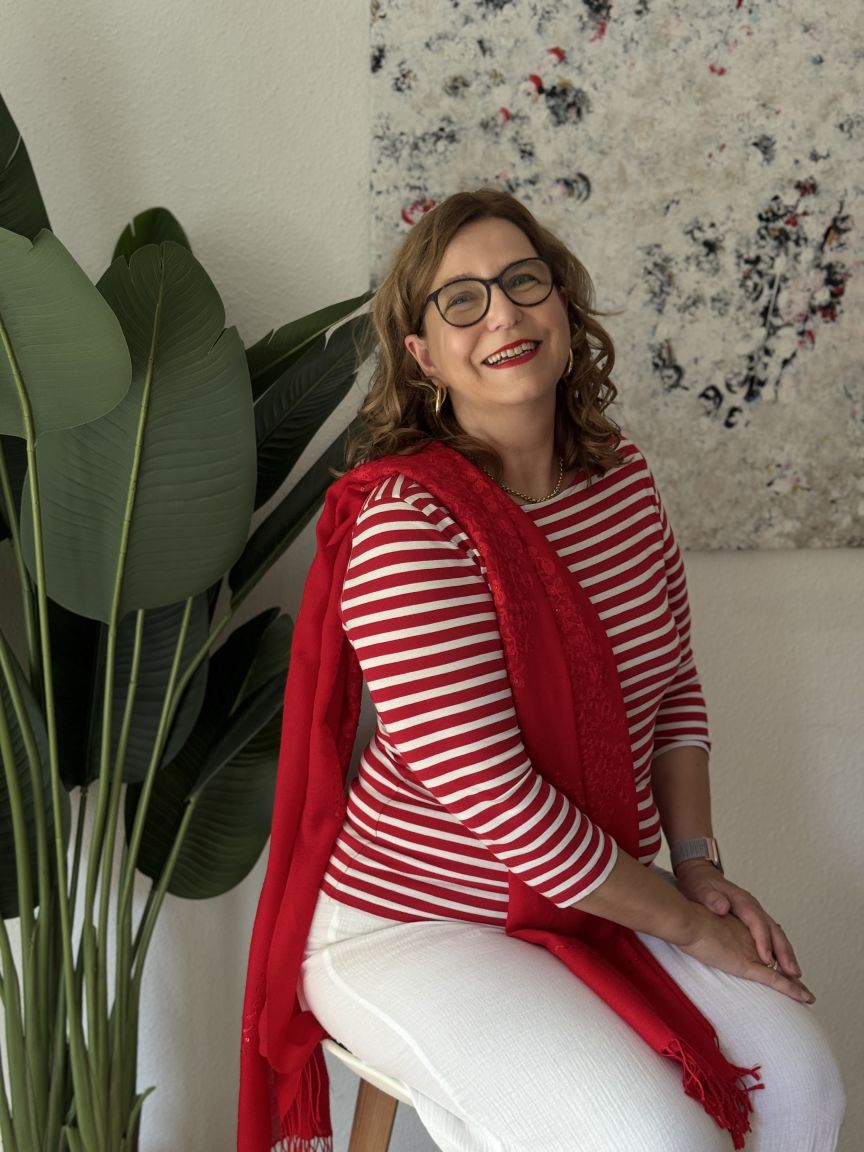 Frau mit Brille, rot-weißem Streifenoberteil und rotem Schal sitzt lächelnd vor Pflanze und abstraktem Bild.