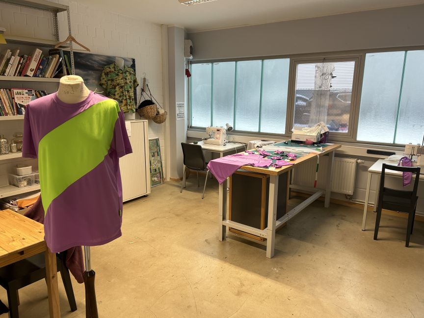 Nähwerkstatt mit Schneiderpuppe in lila-grünem T-Shirt, Arbeitstischen, Nähmaschinen und Regalen mit Büchern.