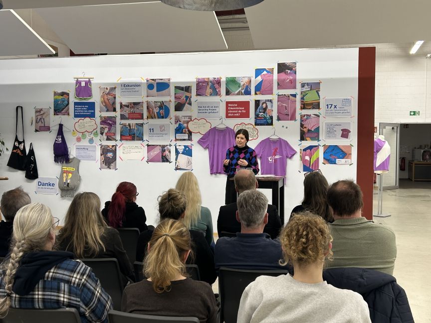 Eine Person steht vor einer Pinnwand mit Fotos und lila T-Shirts und spricht vor einem sitzenden Publikum.
