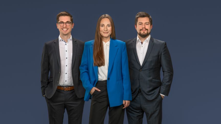 Drei Personen in dunklen Businessanzügen vor blauem Hintergrund; die mittlere Person trägt ein blaues Jacket.
