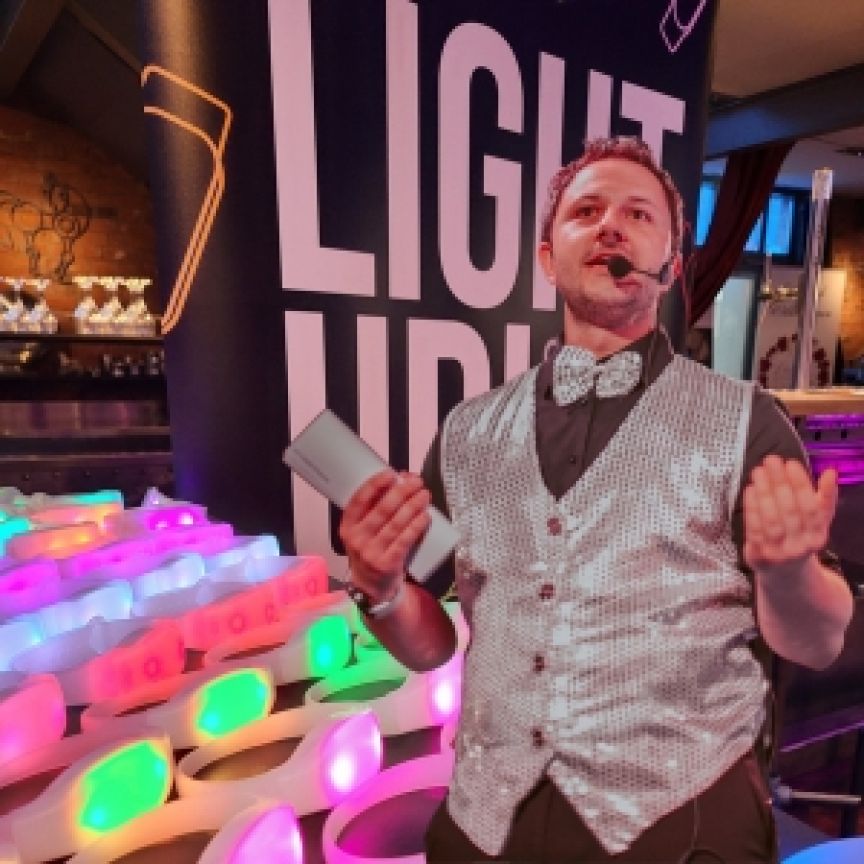 Mann mit Headset-Mikrofon und Fliege präsentiert bunte leuchtende Objekte vor einem "Light Up"-Banner.