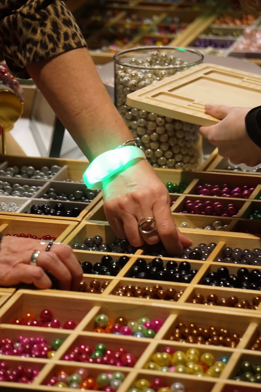 Zwei Hände greifen in Holzboxen mit bunten Perlen. Eine Hand trägt ein leuchtendes grünes Armband und einen Ring.