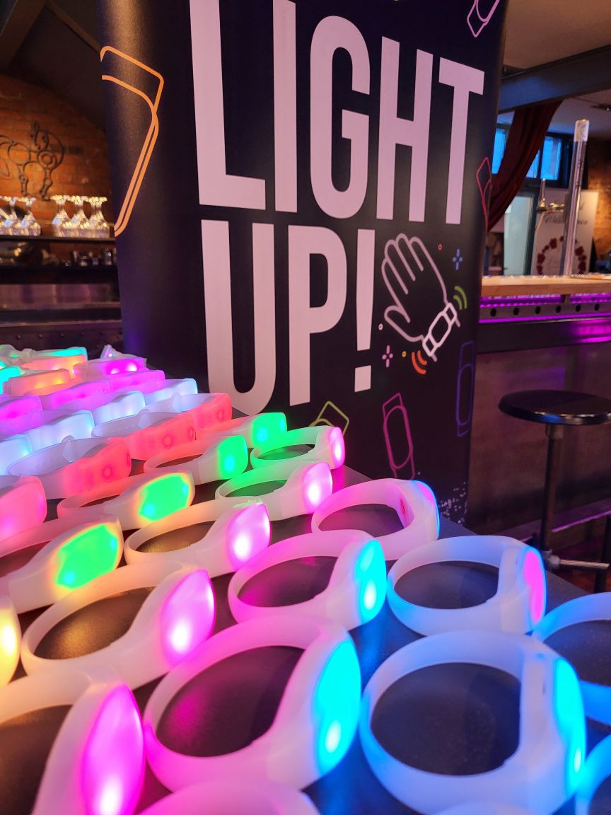 Leuchtende LED-Armbänder in Pink, Grün und Blau vor einem "Light Up!"-Banner in einem Veranstaltungsraum.