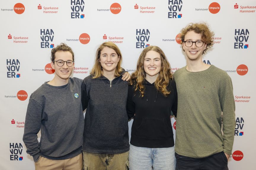 Vier junge Personen stehen lächelnd vor einer Sponsorenwand mit Logos von Sparkasse Hannover und hannover impuls.