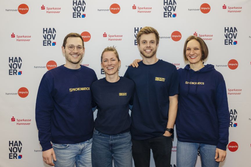 Vier Personen in dunkelblauen "Phonomatics"-Sweatshirts stehen lächelnd vor einer Sparkasse-Hannover-Wand.